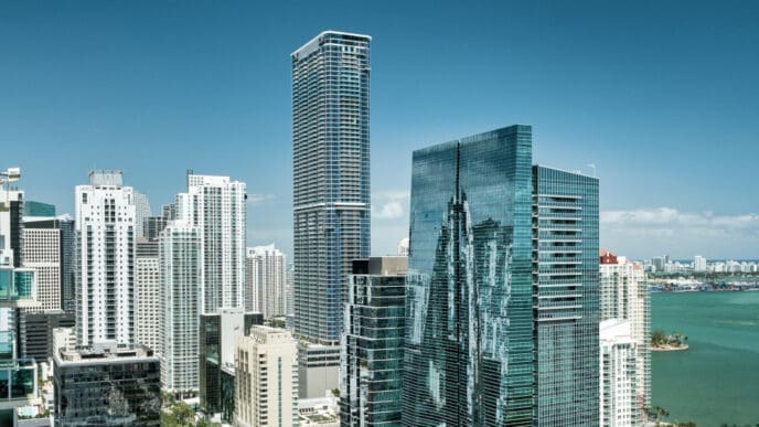 An aerial view of the dense, modern Miami skyline, with numerous tall skyscrapers of glass and white facades, overlooking the blue-green bay with distant land and a port, all under a clear blue sky.
