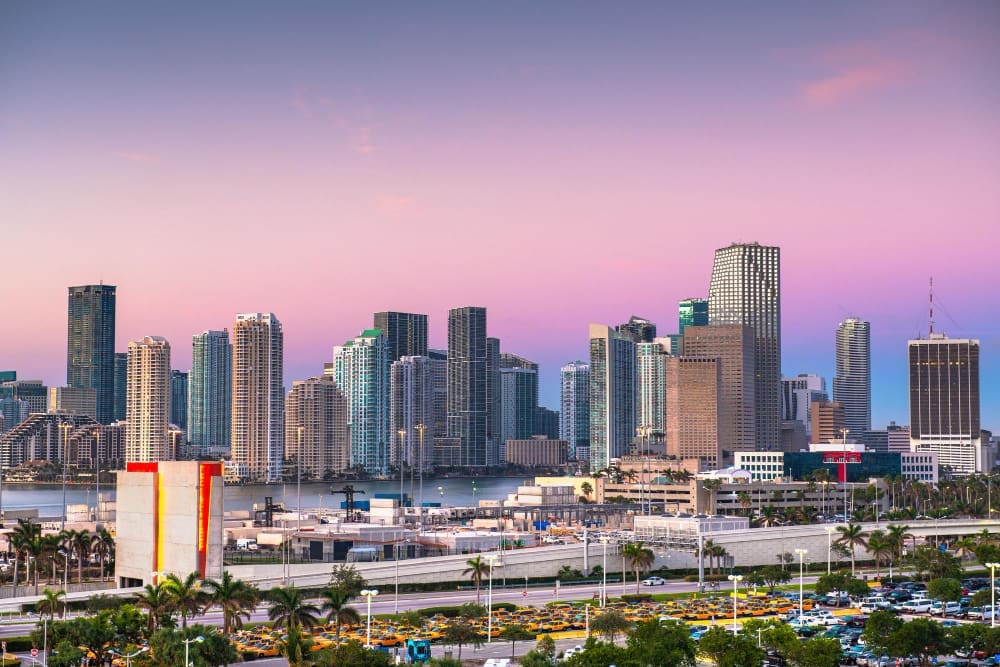 The downtown Miami skyline at sunset, with numerous skyscrapers and high-rises reflecting the soft pink and purple hues of the sky, overlooking a bay and a busy foreground of roads and parking lots.