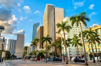 A wide view of downtown Miami, with tall skyscrapers and palm trees lining the wide, multi-lane Biscayne Boulevard on a sunny day.