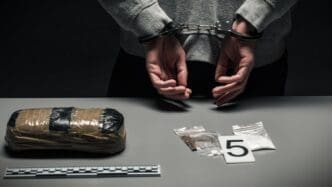 A person is handcuffed behind their back, with a large package of drugs and evidence on a table in front of them