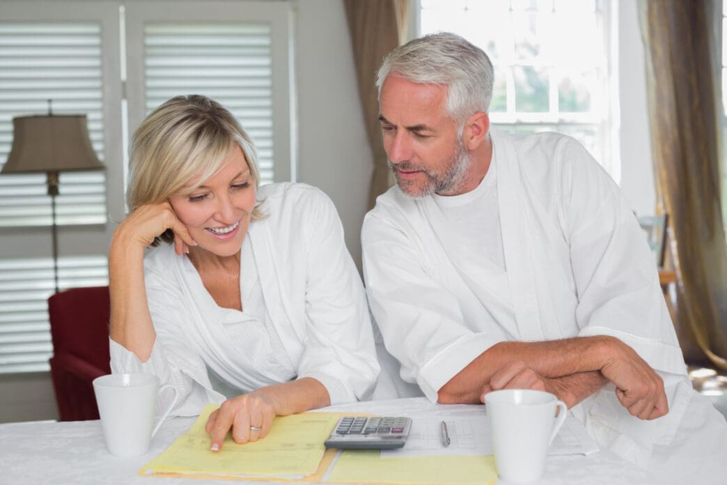 A happy mature couple, both in white robes, sits at a table and reviews financial documents with a calculator.