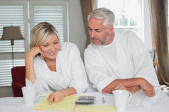 A happy mature couple, both in white robes, sits at a table and reviews financial documents with a calculator.