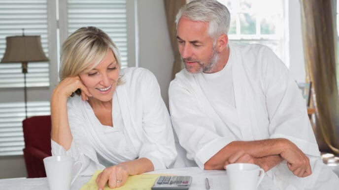 A happy mature couple, both in white robes, sits at a table and reviews financial documents with a calculator.