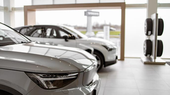 The front of a white electric vehicle is shown in a showroom, with a second car and a tire display in the background.