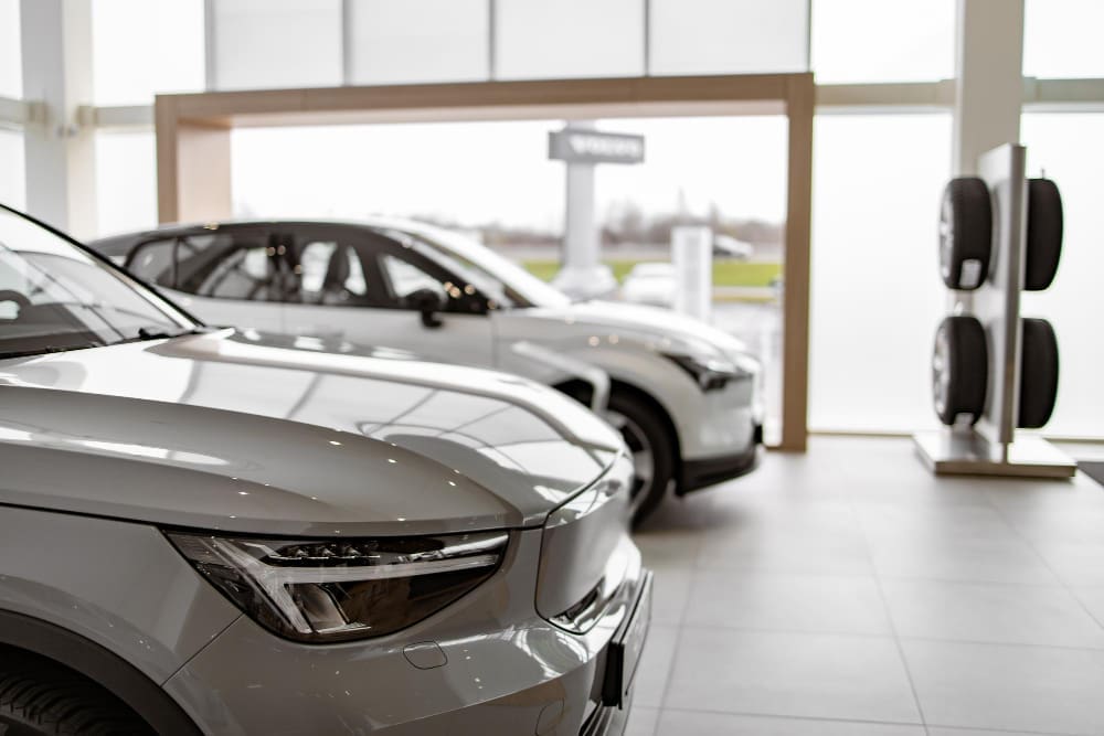 The front of a white electric vehicle is shown in a showroom, with a second car and a tire display in the background.