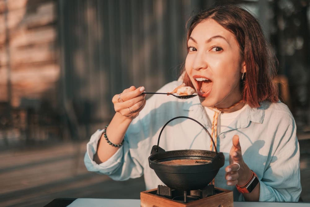 A woman with a joyful expression eating a spoonful of food from a small, black cast-iron pot in an outdoor setting.