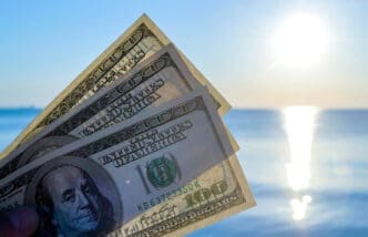 A hand holds up several US hundred-dollar bills against a bright, sun-drenched ocean and sky background.