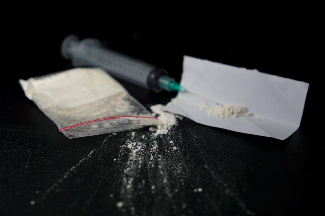 A close-up, dark photograph showing a plastic bag of white powder, a syringe, and a small pile of the powder on a piece of paper.