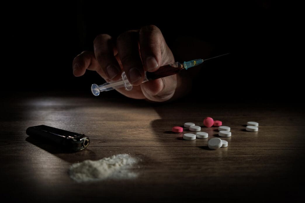 A hand holds a syringe filled with a dark liquid over a table with a pile of white powder and pills