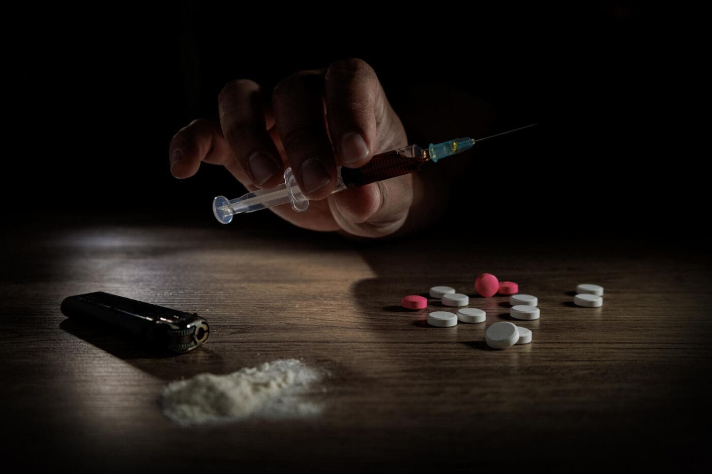 A hand holds a syringe filled with a dark liquid over a table with a pile of white powder and pills