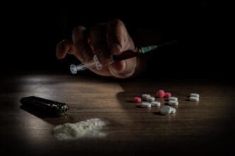 A hand holds a syringe filled with a dark liquid over a table with a pile of white powder and pills