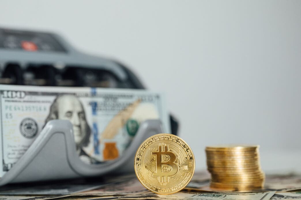 A golden Bitcoin coin and a stack of gold coins are placed on a table, with a blurry money counting machine and a hundred-dollar bill in the background.