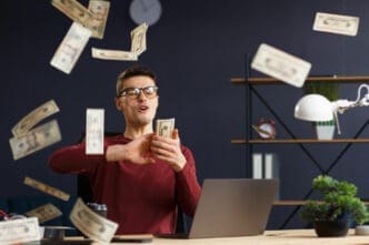 A man with glasses and a red shirt sits at a desk with a laptop, exultantly holding a wad of cash as dollar bills rain down around him, symbolizing financial freedom and passive income.