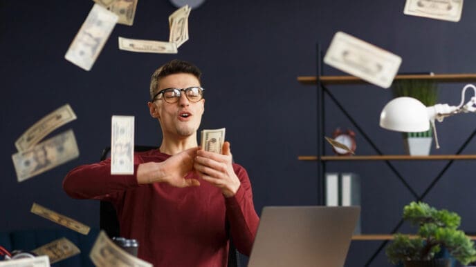 A man with glasses and a red shirt sits at a desk with a laptop, exultantly holding a wad of cash as dollar bills rain down around him, symbolizing financial freedom and passive income.