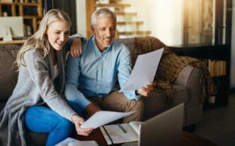 A smiling younger woman and an older man sit on a couch, looking at documents and a laptop.