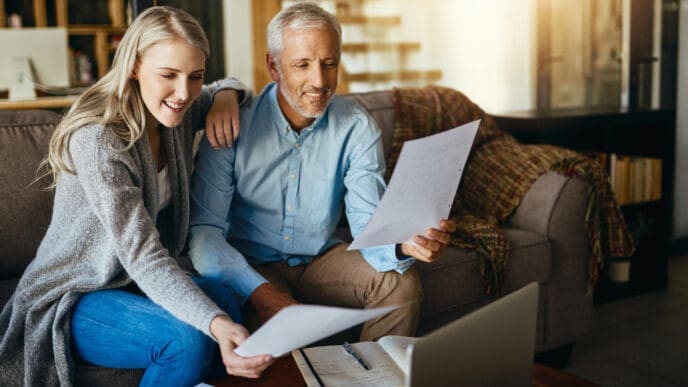 A smiling younger woman and an older man sit on a couch, looking at documents and a laptop.