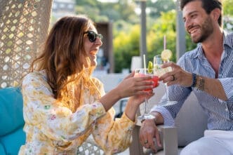A happy couple, a man and a woman, smiles at each other while toasting with colorful cocktails on a sunlit patio.