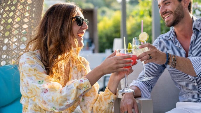 A happy couple, a man and a woman, smiles at each other while toasting with colorful cocktails on a sunlit patio.