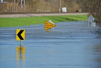 A flooded road with a yellow "NO PASSING ZONE" sign and a left-turn arrow sign partially submerged in the water.