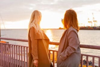 Two women in coats are standing on a pier or dock, looking out at a sunset over the water, with one of them pointing towards the distance.