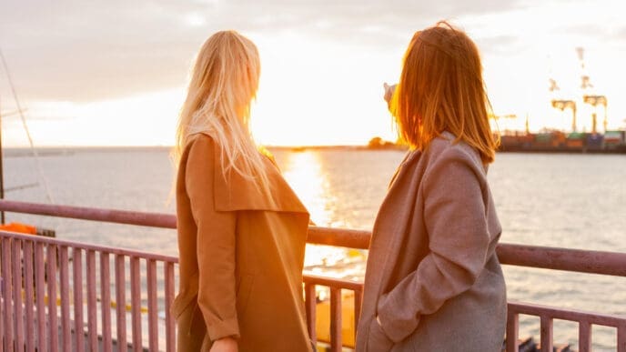 Two women in coats are standing on a pier or dock, looking out at a sunset over the water, with one of them pointing towards the distance.