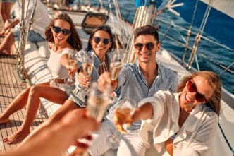 Four young friends on a sailboat are smiling and toasting with champagne glasses at the camera on a sunny day.