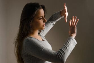 A young woman with long dark hair, wearing a grey long-sleeved shirt, stands in a dark room with her hands raised as if to push something away, her expression showing determination or frustration.