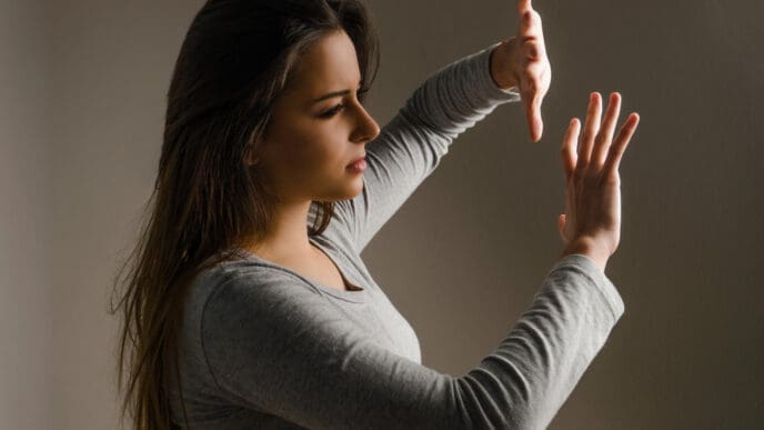A young woman with long dark hair, wearing a grey long-sleeved shirt, stands in a dark room with her hands raised as if to push something away, her expression showing determination or frustration.