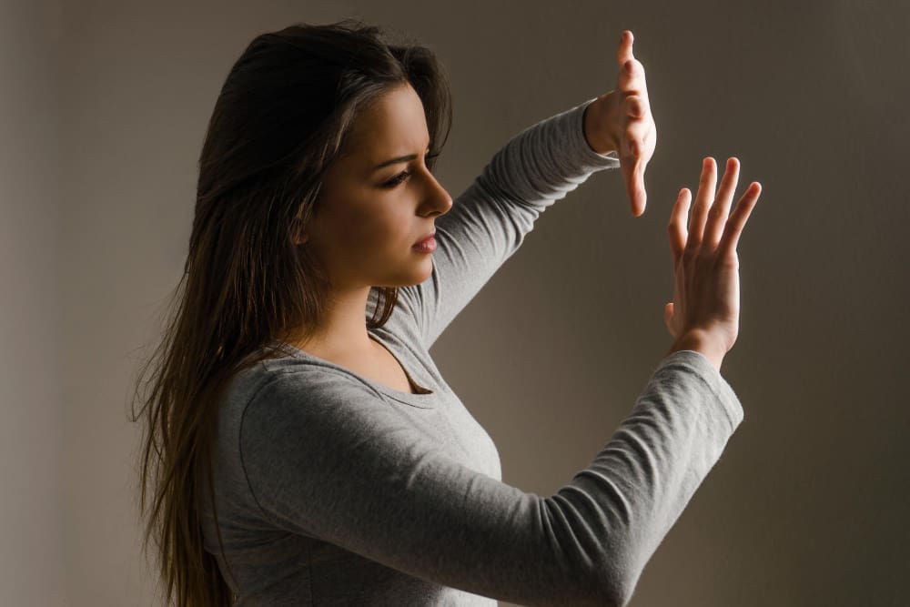 A young woman with long dark hair, wearing a grey long-sleeved shirt, stands in a dark room with her hands raised as if to push something away, her expression showing determination or frustration.