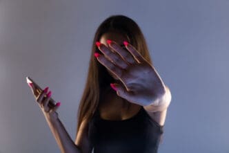 A person holds a hand up to block their face from view, while holding a smartphone in the other hand.