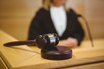 A judge's gavel is on a wooden bench, with the blurry figure of a judge in the background.