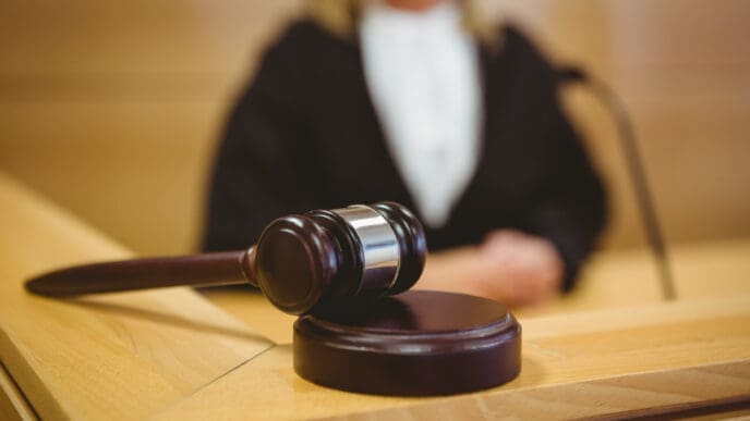 A judge's gavel is on a wooden bench, with the blurry figure of a judge in the background.