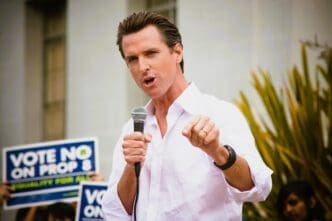 Gavin Newsom, a man in a white collared shirt, holds a microphone and points at a crowd while a person behind him holds a sign that says "VOTE NO ON PROP 8".