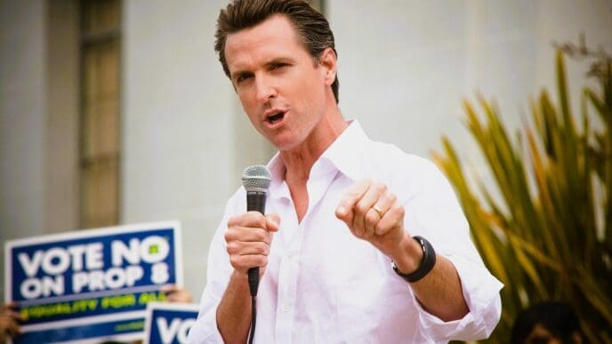 Gavin Newsom, a man in a white collared shirt, holds a microphone and points at a crowd while a person behind him holds a sign that says "VOTE NO ON PROP 8".