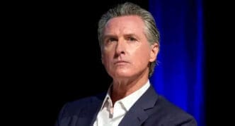 A close-up portrait of Governor of California Gavin Newsom, with a serious expression and graying hair, wearing a suit.