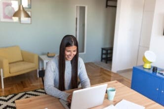 A smiling woman with long dark hair sits at a wooden desk and works on her laptop in a sunlit modern home office.