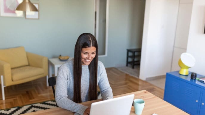 A smiling woman with long dark hair sits at a wooden desk and works on her laptop in a sunlit modern home office.