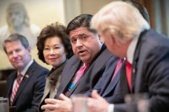 President Donald Trump is seated at a table, with his back to the camera, listening to J.B. Pritzker who is speaking.