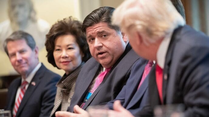 President Donald Trump is seated at a table, with his back to the camera, listening to J.B. Pritzker who is speaking.