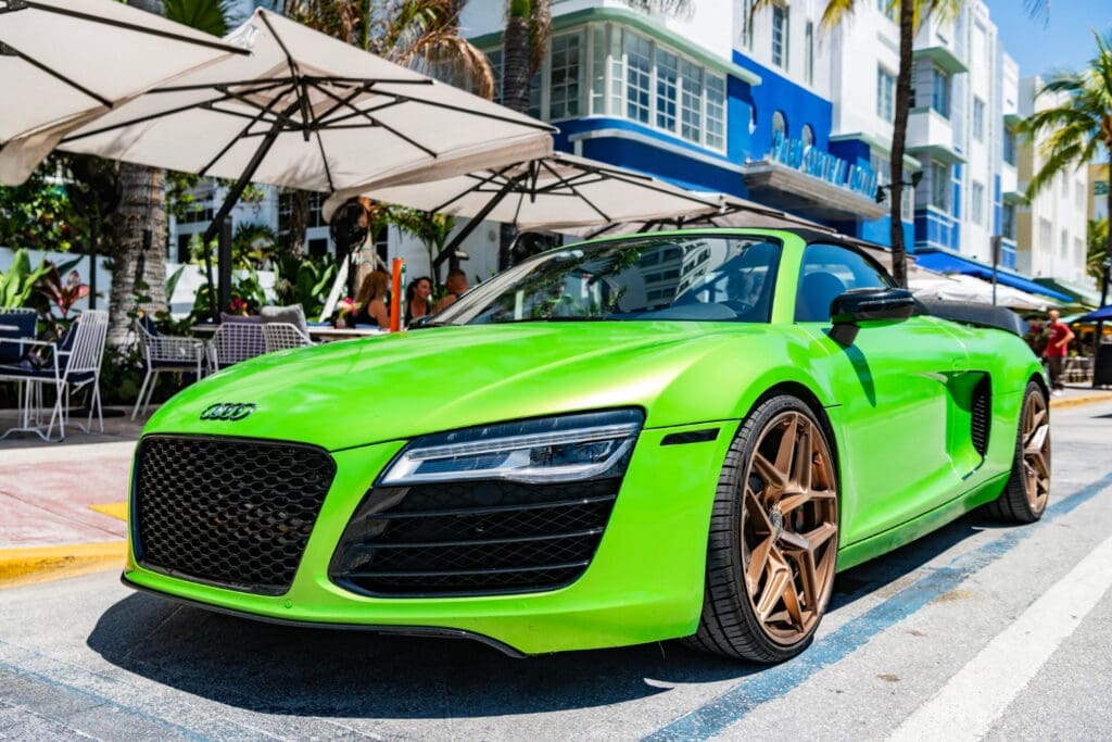 A vibrant, fluorescent green Audi R8 convertible sports car with bronze rims is parked on a street in Miami Beach in front of a sidewalk cafe and art deco hotel.