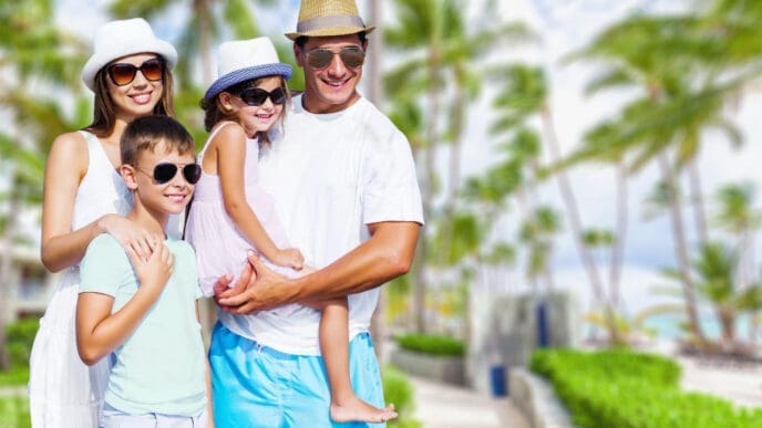 A happy family of four in sunglasses and hats, posing for a portrait on a sunny day in Miami Beach with palm trees in the background.