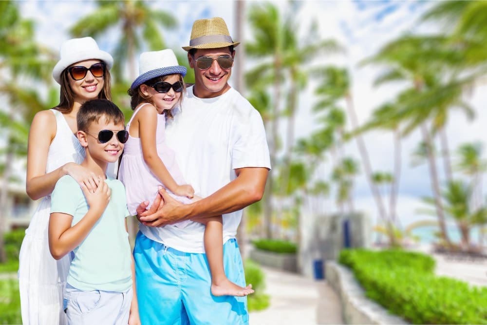 A happy family of four in sunglasses and hats, posing for a portrait on a sunny day in Miami Beach with palm trees in the background.