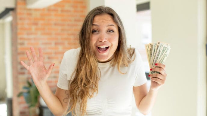 A young woman with an astonished expression holds a fan of U.S. dollar bills in one hand and has her other hand raised.