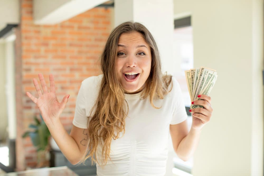A young woman with an astonished expression holds a fan of U.S. dollar bills in one hand and has her other hand raised.