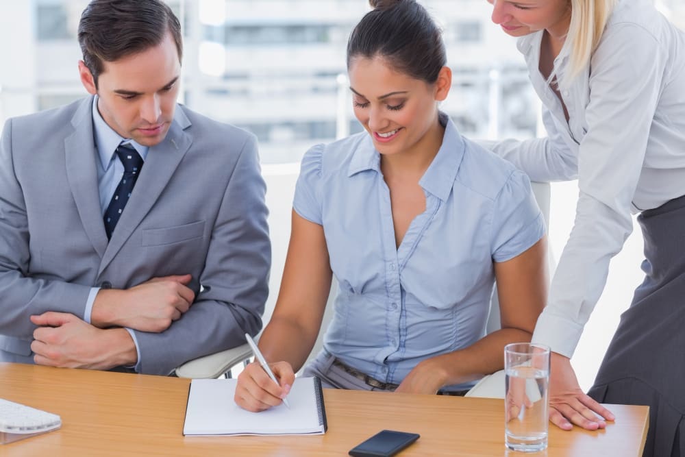 Three businesspeople in an office, with two of them looking at notes on a notepad while the third offers advice.