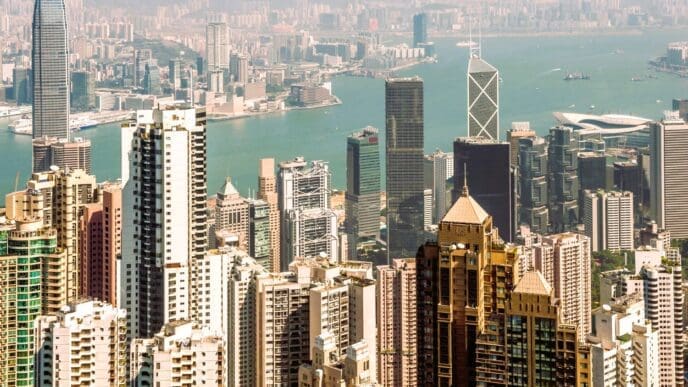 An aerial view of the incredibly dense, high-rise urban landscape of Hong Kong, with Victoria Harbour and mountains in the distance.