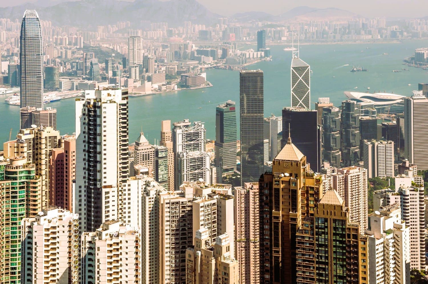 An aerial view of the incredibly dense, high-rise urban landscape of Hong Kong, with Victoria Harbour and mountains in the distance.