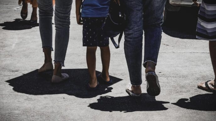 A low-angle shot of a group of people walking on a sun-drenched road, with strong shadows cast on the ground.