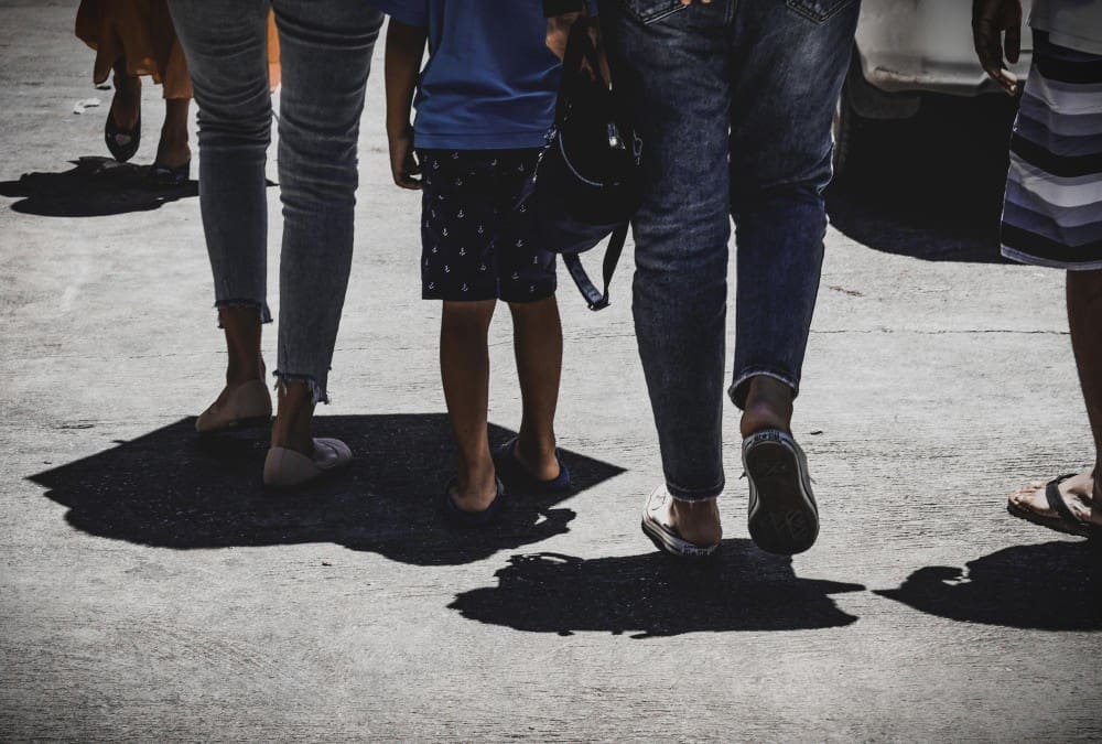 A low-angle shot of a group of people walking on a sun-drenched road, with strong shadows cast on the ground.