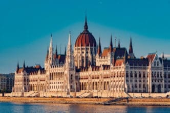 A full view of the Hungarian Parliament Building, a large Gothic Revival structure with a central dome and many spires, standing on the bank of a river under a clear blue sky.